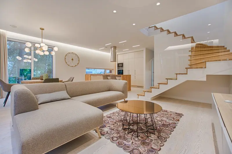 Living Room Image With Light Brown or Caranek color sofa chairs, Gold table at the center, and stairs on the right side with Kitchen on the back