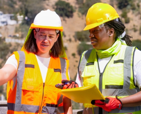 Image of two women wearing safety helmet and uniform, the woman on the left side holding a paper and seems like they're talking about something.