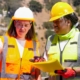 Image of two women wearing safety helmet and uniform, the woman on the left side holding a paper and seems like they're talking about something.