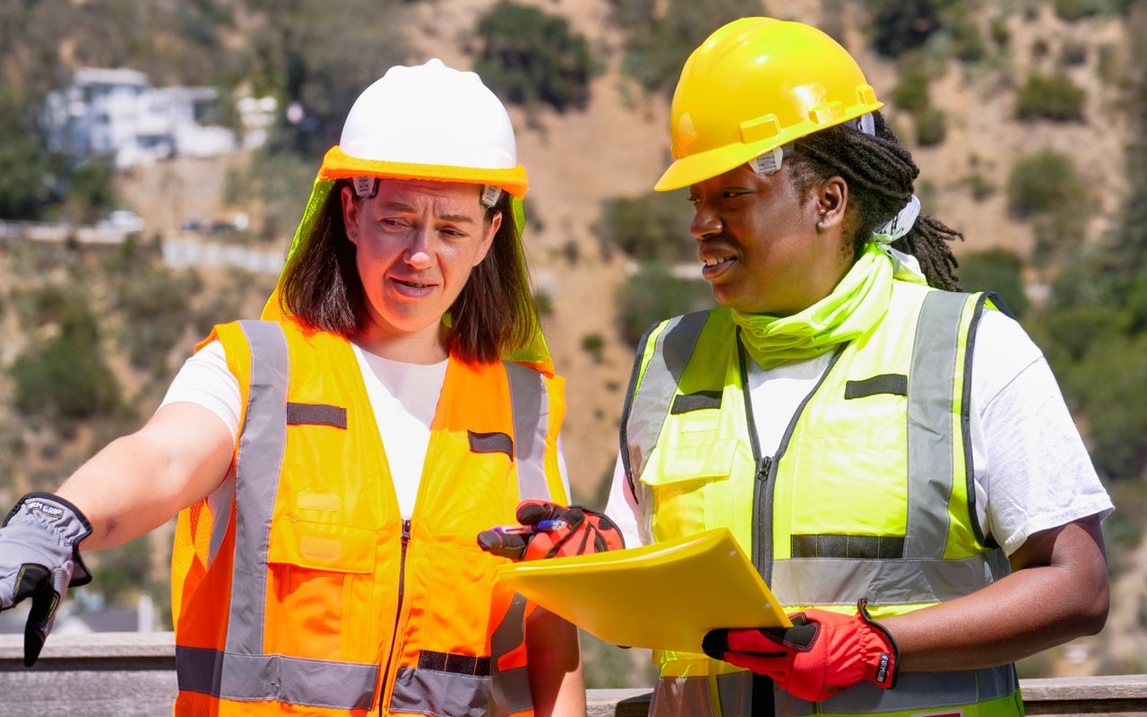 Image of two women wearing safety helmet and uniform, the woman on the left side holding a paper and seems like they're talking about something.